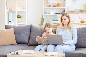 Portrait of adult working mother using laptop while sitting on couch at home with cute daughter reading book beside her, copy space
